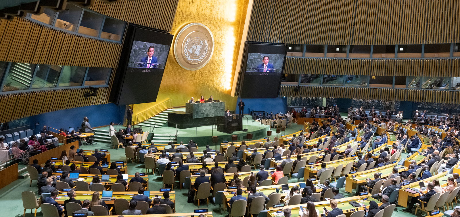 The United Nations General Assembly hall in session, delegates at their desks below the gold emblem