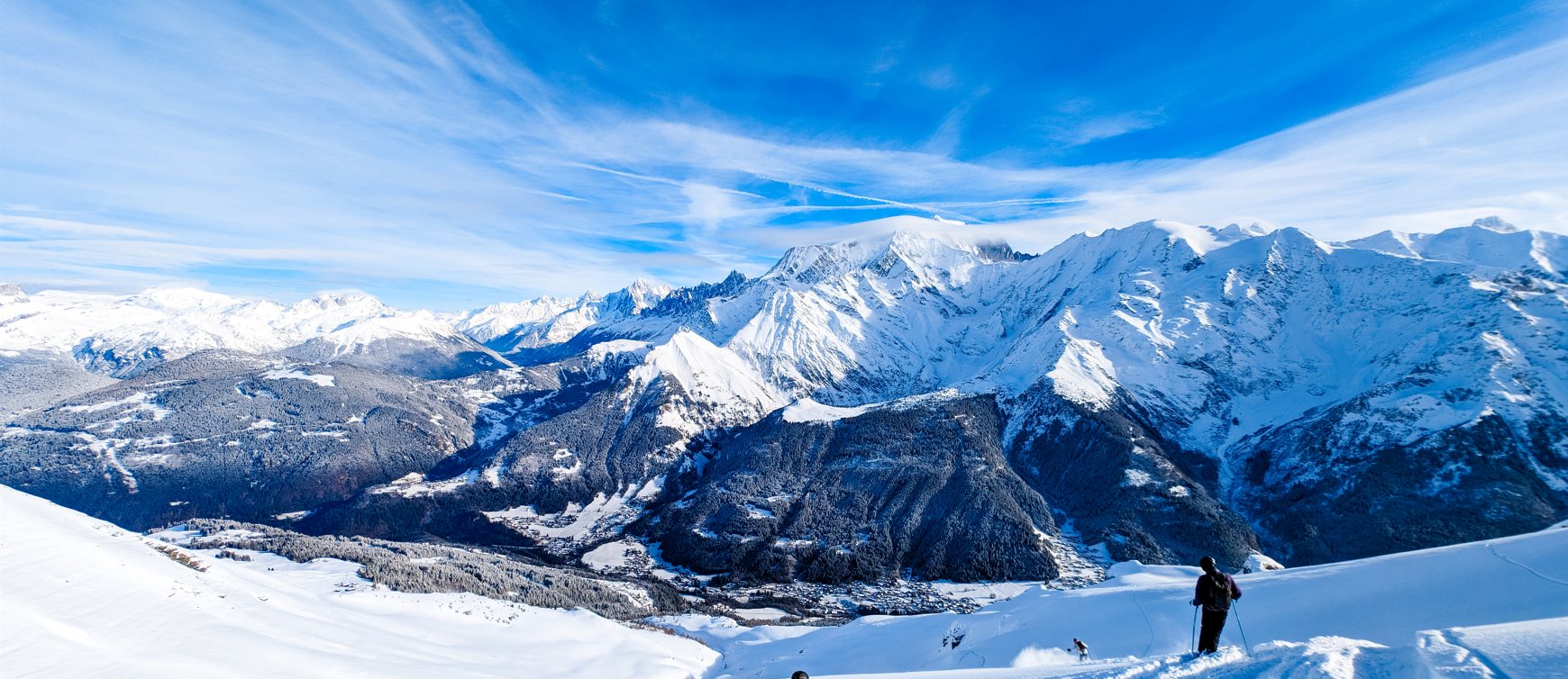 Snow covered Alpine peaks under a clear blue sky, a forested valley with a village far below, and a lone skier standing on the near slope